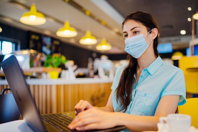 Woman in coffee shop working on laptop in mask